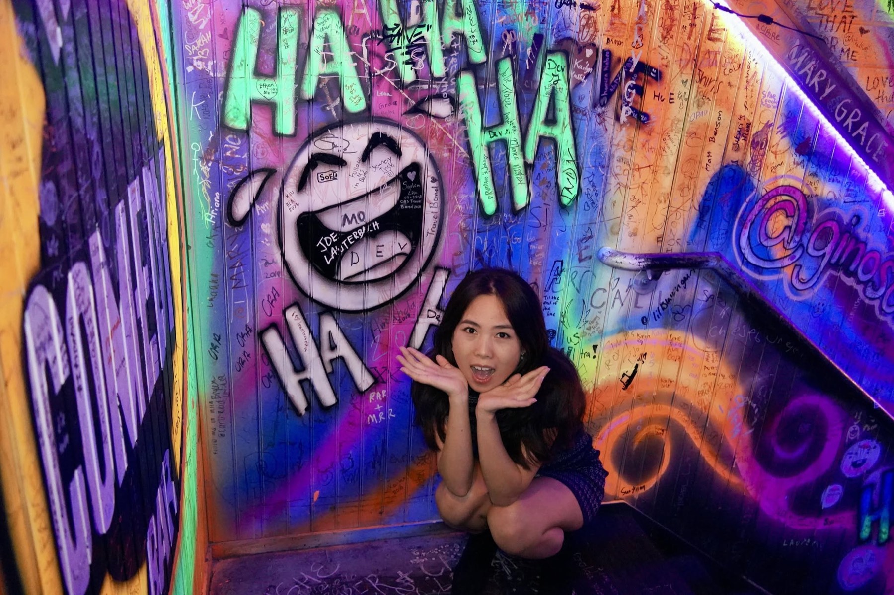 Yaqiao Yang crouched in a neon graffiti stairwell covered in HA HA murals, hands under her chin