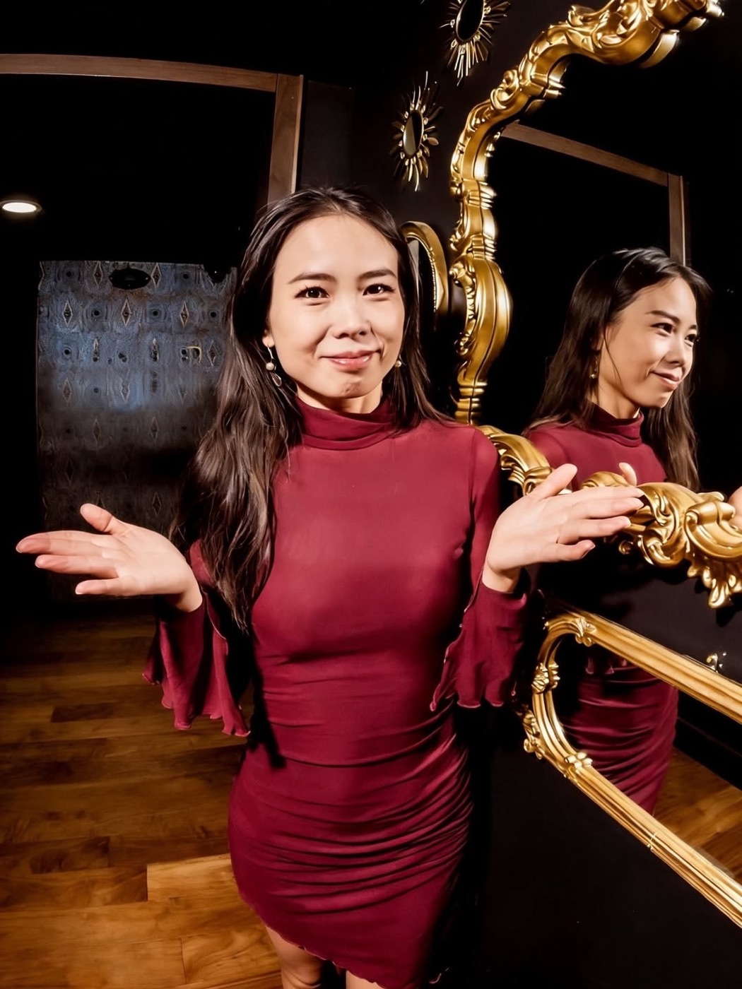 Yaqiao Yang in a burgundy dress, shrugging beside a gilded mirror
